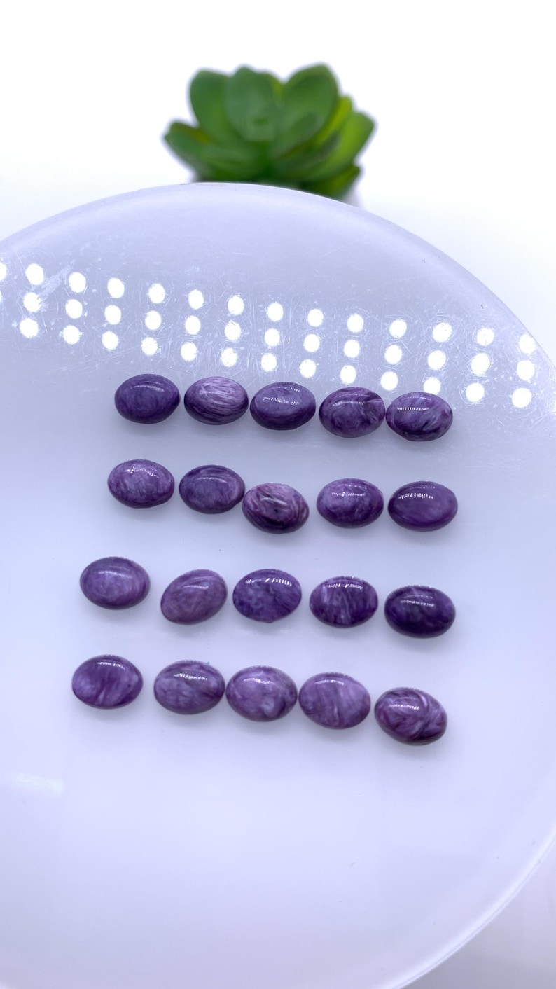 May include: A collection of 18 oval-shaped purple gemstones, arranged in rows on a white surface.