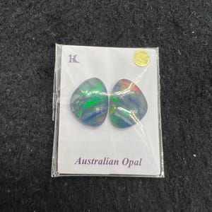 May include: Two Australian opal cabochons in a clear plastic package with a white card that says "Australian Opal". The opals are a dark blue-green color with flashes of red and green.