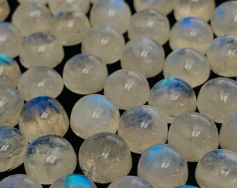 Op de afbeelding: Een verzameling ronde, witte maansteen cabochons met iriserende blauwe flitsen. De stenen zijn glad en gepolijst en zijn in een willekeurig patroon gerangschikt.