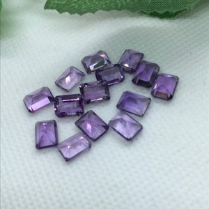 May include: A group of 12 purple amethyst gemstones cut in a rectangular emerald shape.