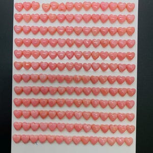 May include: Rows of small, heart-shaped, pink beads arranged on a white surface. The beads are a soft, pastel pink color and appear to be made of a smooth, glossy material. The image includes the text "168 pcs".