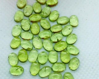 Puede incluir: Un conjunto de gemas de peridoto ovaladas y translúcidas. Las piedras son de un color verde claro vibrante con inclusiones visibles. Las gemas están dispersas sobre una superficie blanca, con unos pétalos rosas en la esquina superior izquierda.