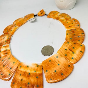 May include: A necklace made of large, rectangular, orange and white bone beads with turquoise accents. The beads are arranged in a curved shape, and the necklace is strung on a black cord.