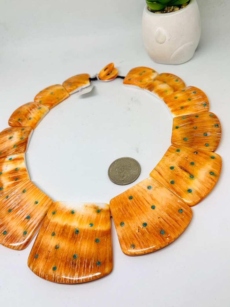 May include: A necklace made of large, rectangular, orange and white bone beads with turquoise accents. The beads are arranged in a curved shape, and the necklace is strung on a black cord.