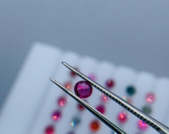 May include: A close-up of a small, round, faceted, deep red gemstone held by silver tweezers. Other gemstones of various colors are visible in the background, scattered on a white surface. The lighting is bright and the background is blurred.