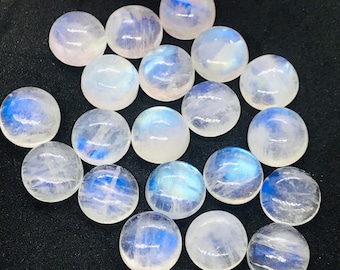 Puede incluir: Una colección de 18 cabujones redondos de piedra lunar blanca con destellos iridiscentes azules y blancos. Los cabujones son lisos y pulidos, y están dispuestos en un patrón aleatorio sobre una superficie negra.