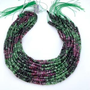 May include: Strands of faceted gemstone beads in shades of green, dark green, and magenta. The beads are strung together with green thread, and the strands are arranged in a curved shape against a white background. The beads appear to be small and square.