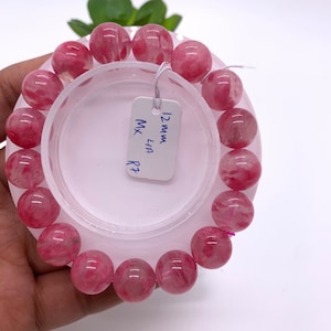 May include: A pink quartz bracelet with 12mm beads. The bracelet is on a white display stand with a tag that reads "12mm MX LIA R7".
