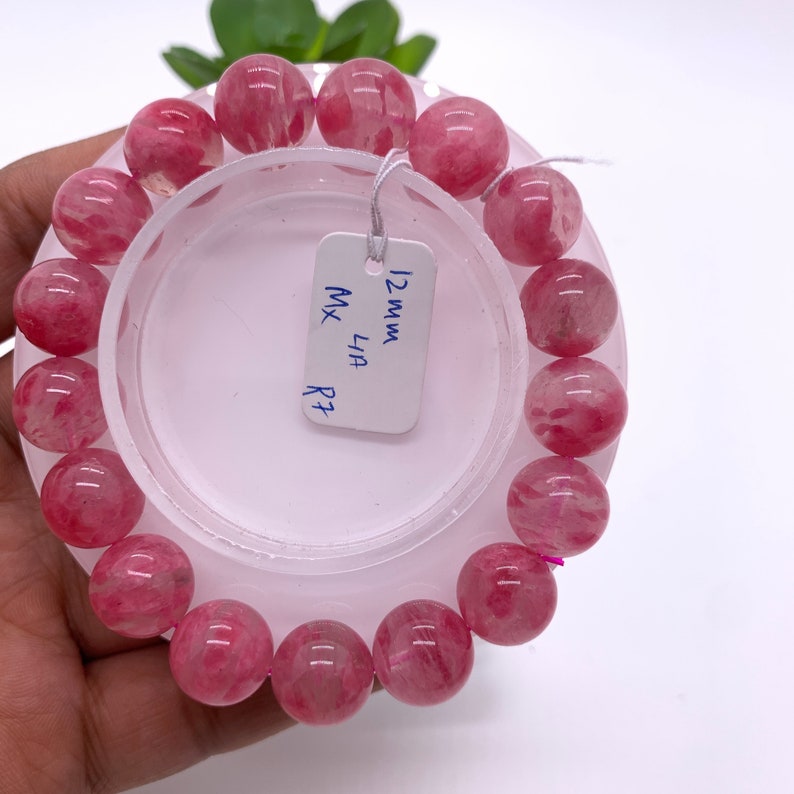 May include: A pink quartz bracelet with 12mm beads. The bracelet is on a white display stand with a tag that reads "12mm MX LIA R7".