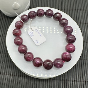 May include: A purple gemstone bracelet with 12 mm beads. The bracelet is on a white plate with a tag that says "Rex CT 12 mm".
