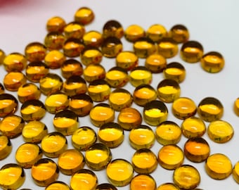 May include: A large group of round, amber-colored glass cabochons. The cabochons are scattered on a white surface.
