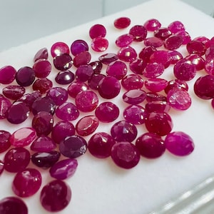 May include: A collection of loose, round, faceted ruby gemstones in various shades of pink and red. The gemstones are scattered on a white surface.