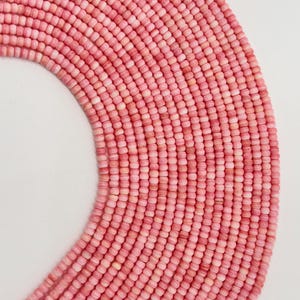 Puede incluir: Primer plano de una colección de cuentas rosas. Las cuentas son pequeñas, cilíndricas y están dispuestas en filas ordenadas, creando una superficie texturizada. El color varía de rosa claro a medio, con algunas cuentas que parecen casi blancas. Las cuentas son probablemente para hacer joyas.