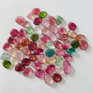 May include: A collection of polished gemstones in various shades of pink, red, green, and clear. The stones are oval-shaped and scattered on a white surface. The colors range from deep magenta to pale pink and translucent green.