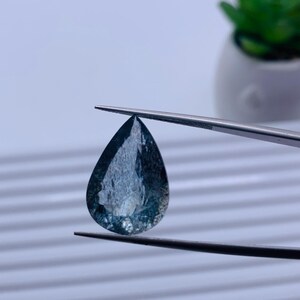 May include: A pear-shaped gemstone with a blue-green hue and white inclusions. The gemstone is held by tweezers against a white background.