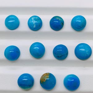 May include: Ten round, blue turquoise cabochons, arranged in three rows on a white surface.