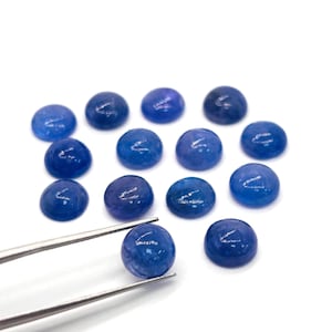 May include: A collection of round, polished blue gemstones. One gemstone is held by silver tweezers, showcasing its smooth, domed surface. The other gemstones are scattered on a white background, displaying variations in color and depth.