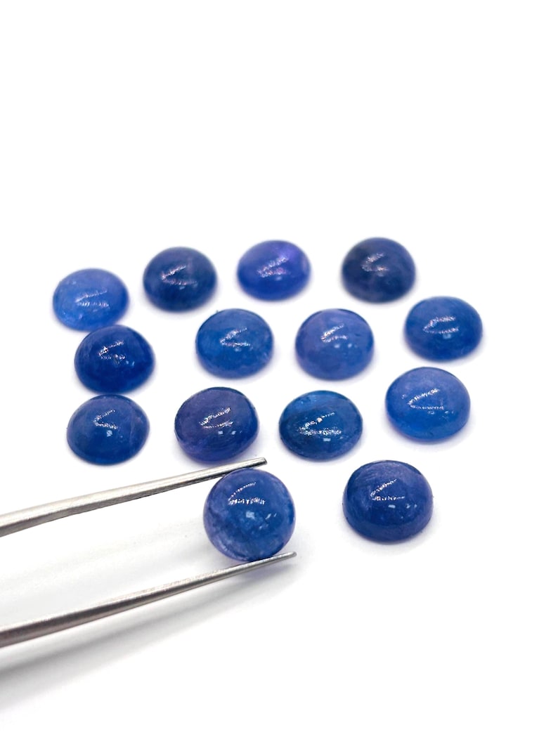 May include: A collection of round, polished blue gemstones. One gemstone is held by silver tweezers, showcasing its smooth, domed surface. The other gemstones are scattered on a white background, displaying variations in color and depth.