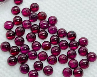 May include: A collection of small, round, polished gemstones in varying shades of deep red and purple. The gemstones are scattered across a white surface, with some appearing to be in a clear container. The gems have a smooth, reflective surface.