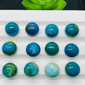 May include: Twelve round, blue and green gemstone cabochons arranged in three rows of four on a white surface. The cabochons have a variety of patterns and textures.