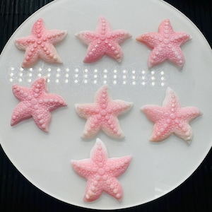 May include: Seven pink and white starfish-shaped soaps on a white plate. The soaps are arranged in a circle.
