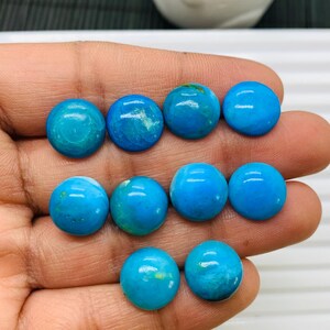 Può includere: Dieci cabochon rotondi in turchese, ognuno con un motivo leggermente diverso di vortici blu e bianchi. I cabochon sono disposti in due file su una mano.