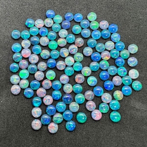 Puede incluir: Una colección de cabujones de ópalo redondos e iridiscentes en tonos azules, verdes y rosas. Los cabujones tienen un diámetro aproximado de 6 mm y un acabado liso y pulido.