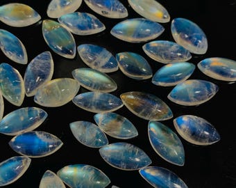 Puede incluir: Una colección de piedras preciosas de piedra lunar en forma ovalada. Las piedras muestran una gama de colores, incluyendo azules, amarillos y blancos. Las piedras preciosas están pulidas y tienen una superficie lisa y reflectante. Las piedras están dispersas sobre una superficie oscura.