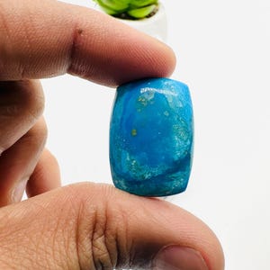 May include: A bright blue cabochon gemstone with a rectangular shape and a slightly irregular surface. The stone has a mottled appearance with hints of green and white.