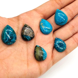 May include: A collection of teardrop-shaped gemstones in shades of turquoise and brown. The stones are held in a hand, showcasing their natural textures and variations. The gemstones are likely used for jewelry or crafting.