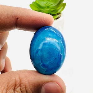 May include: A blue and white oval cabochon gemstone with a swirling pattern. The gemstone is being held by a hand.