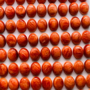 May include: A collection of oval-shaped, polished gemstones in shades of orange and brown, arranged in rows. The gemstones have a marbled appearance. The dimensions are 8x10x4mm. The image is taken from a high angle.