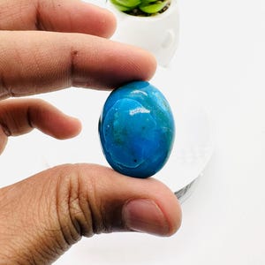 May include: A smooth, oval-shaped turquoise cabochon gemstone with a blue and green color pattern. The stone is held in a hand.