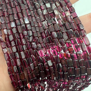 May include: A strand of dark red gemstone beads, each bead is a rectangular prism with slightly rounded edges. The beads are strung together to form a long necklace.