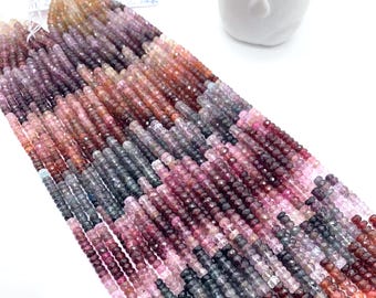 May include: Strands of faceted gemstone beads in shades of pink, purple, gray, and brown. The beads are small and uniform in size, strung together to create a flowing, textured appearance. A small, green succulent plant in a white pot is in the background.