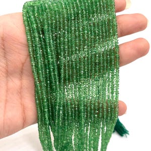 May include: Strands of small, round, bright green beads. The beads are strung together, forming multiple strands. The beads have a slightly translucent appearance. The background is white. The beads are held in a hand.