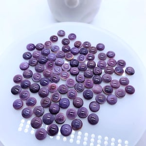 May include: A collection of small, round, purple buttons with a glossy finish, scattered on a white plate. The buttons have a subtle, layered design and varying shades of purple. A white ceramic object is in the background.