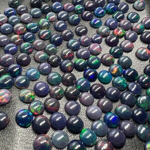 Può includere: Una collezione di cabochon di opale nero con colori arcobaleno iridescenti. Gli opali sono disposti a formare un motivo su una superficie grigio scuro.