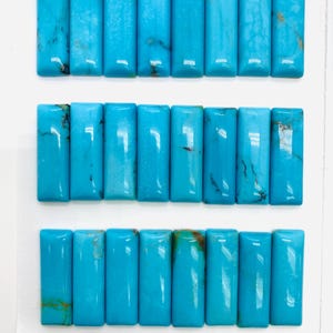 May include: A collection of 24 rectangular turquoise gemstones, each approximately 8mm x 24mm, arranged in three rows on a white surface. The gemstones are a vibrant blue with natural veining and variations in color. The text "8 x 24mm" and "24pcs" is visible.