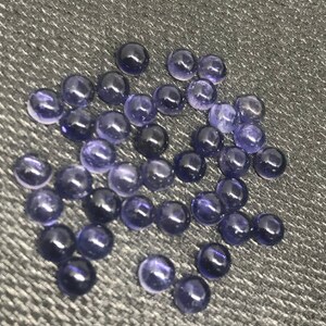 May include: A collection of approximately 40 small, round, purple gemstones. The gemstones are scattered on a dark gray textured surface.