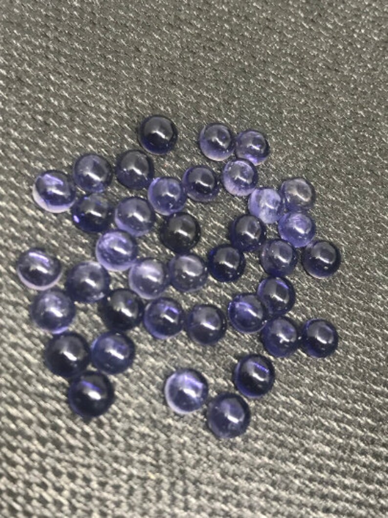 May include: A collection of approximately 40 small, round, purple gemstones. The gemstones are scattered on a dark gray textured surface.