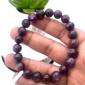 Puede incluir: Una pulsera de cuentas compuesta por cuentas redondas de piedra preciosa pulida de color púrpura. Las cuentas muestran tonos variados de violeta intenso y ciruela con vetas naturales internas y una superficie lisa y reflectante, ensartadas en un cordón elástico.