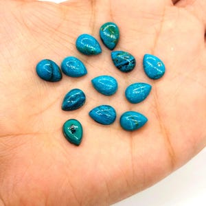 May include: A collection of teardrop-shaped turquoise gemstones, each displaying a vibrant blue hue with natural veining. The polished stones vary slightly in color and pattern, showcasing the unique characteristics of natural turquoise. These gemstones are ideal for jewelry making.