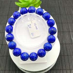Puede incluir: Un brazalete de lapislázuli azul con cuentas de 12 mm. El brazalete está en un expositor blanco con una etiqueta que dice "12 mm Lapis".