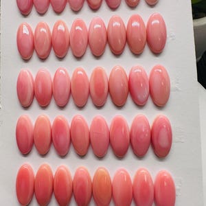 May include: A collection of 40 oval-shaped pink shell cabochons, each measuring 7x15mm, arranged in rows on a white background.