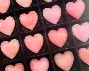Könnte beinhalten: Eine Sammlung von rosa herzförmigen Muschelkabochen, die in einem schwarzen Display-Kasten angeordnet sind. Die Muscheln sind poliert und haben eine glatte, glänzende Oberfläche.