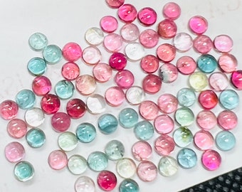 May include: A collection of round, faceted gemstones in various shades of pink, green, and blue. The gemstones are scattered on a white surface.