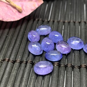 May include: A group of oval-shaped, light blue gemstones with a slight purple hue. The gemstones are arranged on a black, woven surface.