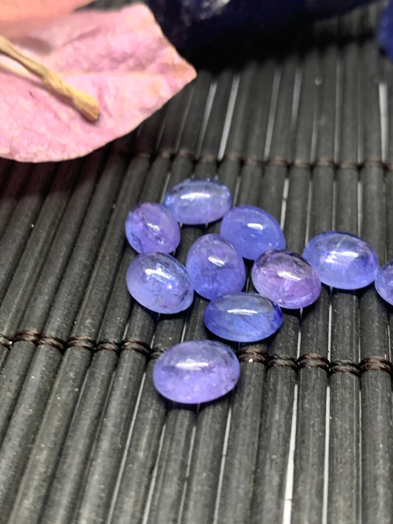 May include: A group of oval-shaped, light blue gemstones with a slight purple hue. The gemstones are arranged on a black, woven surface.
