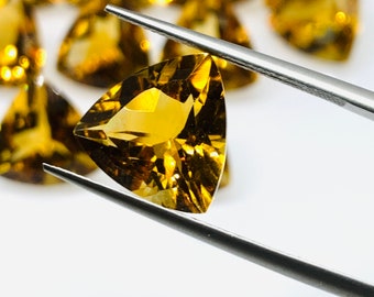 Puede incluir: Primer plano de una piedra preciosa triangular de color amarillo dorado sostenida por unas pinzas. La piedra preciosa está facetada y tiene un alto grado de brillo.